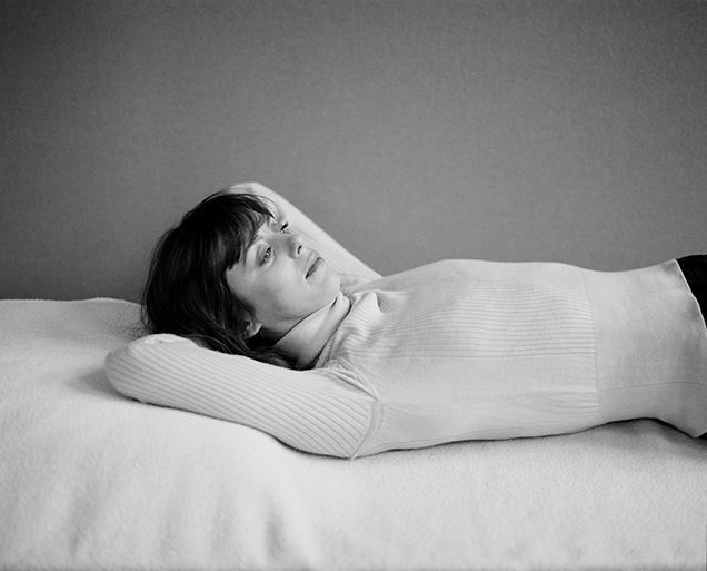 Black and White Photo of Helen Amy Murray reclining with her arms behind her head on a white blanket with a grey background