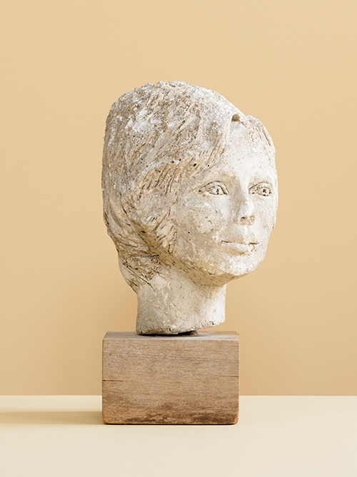 Photograph of a weathered plaster sculpture by Jane Murray, of a female head on a wooden block against a cream background