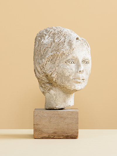 Photograph of a weathered plaster sculpture by Jane Murray, of a female head on a wooden block against a cream background
